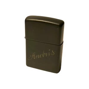 Puede incluir: Un encendedor de metal gris oscuro con el nombre "Andris" grabado en cursiva en la parte delantera. El encendedor tiene forma rectangular con bordes redondeados y una tapa con bisagras.