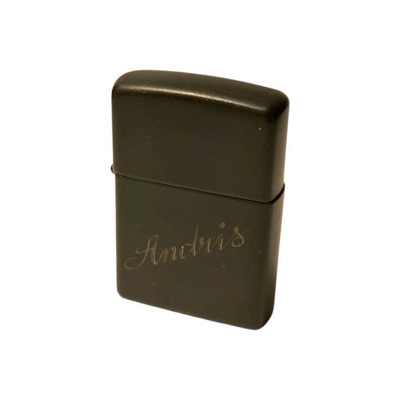 Puede incluir: Un encendedor de metal gris oscuro con el nombre "Andris" grabado en cursiva en la parte delantera. El encendedor tiene forma rectangular con bordes redondeados y una tapa con bisagras.