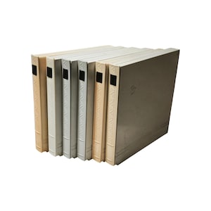 May include: A collection of seven vintage binders in varying shades of cream and off-white. Each binder has a black label holder and a decorative embossed design along the spine. The binders are stacked together.