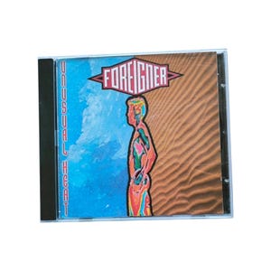 Op de afbeelding: Een cd-hoesje met het album "Unusual Heat" van Foreigner. De cover heeft een gesplitst ontwerp: blauw water aan de linkerkant en een zandachtige textuur aan de rechterkant. De bandnaam staat in rode en witte tekst boven een kleurrijk silhouet.