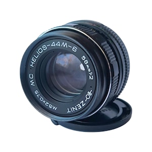 May include: A black and silver Helios-44M-6 58mm f/2 camera lens with a black lens cap. The lens has the text "Helios-44M-6", "58mm 1:2", and "MC" printed on it. The lens is designed for photography.