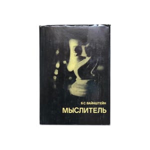 Puede incluir: Un libro de tapa dura con una cubierta negra que presenta un diseño de pieza de ajedrez en amarillo y blanco. El libro tiene texto en ruso, incluido el título "Mыслитель" y el nombre del autor.