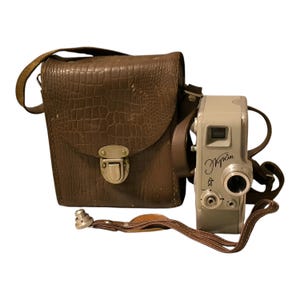 Puede incluir: Una cámara de cine de 8 mm vintage con una funda de cuero marrón. La cámara es plateada con una lente negra y el texto "КРАЙ" escrito en el lateral.