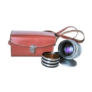 May include: A vintage camera lens with a silver body and a leather carrying case. The lens is accompanied by a set of filters and a lens cap. The case is a reddish-brown color and has a shoulder strap.
