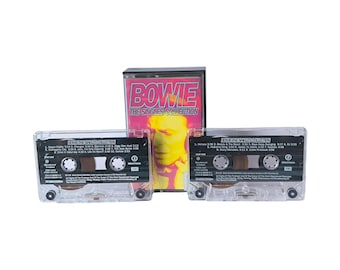 May include: A David Bowie The Singles Collection cassette tape set featuring a clear plastic case with a pink and yellow cover insert. Two transparent cassette tapes are included, each with black labels listing track titles and production details.
