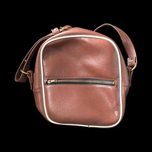 Könnte beinhalten: Eine rechteckige, braune Umhängetasche mit weißem Rand und einem schwarzen Reißverschluss. Die Tasche hat einen oberen Griff und einen Schultergurt. Die Tasche ist aus lederähnlichem Material.