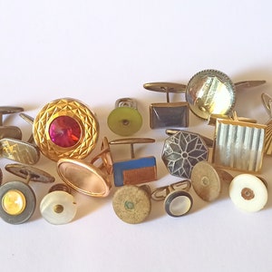 Op de afbeelding: Een verzameling vintage manchetknopen in verschillende stijlen en kleuren, waaronder goud, zilver, zwart, wit en rood. Sommige manchetknopen hebben geometrische ontwerpen, terwijl andere een meer sierlijke uitstraling hebben.