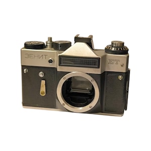 Op de afbeelding: Een vintage zilveren en zwarte camera met de tekst "ЗЕНИТ" aan de bovenkant. De camera heeft een lensopening, een flitsschoen en diverse draaiknoppen en knoppen. De camera is een klassieke filmcamera.