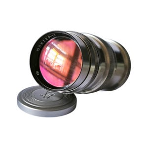 Può includere: Un obiettivo per fotocamera vintage con corpo argentato e tappo. L'obiettivo ha un vetro riflettente con una tinta rosa. Il tappo è argentato e presenta un logo. L'obiettivo è etichettato "Jupiter-11".