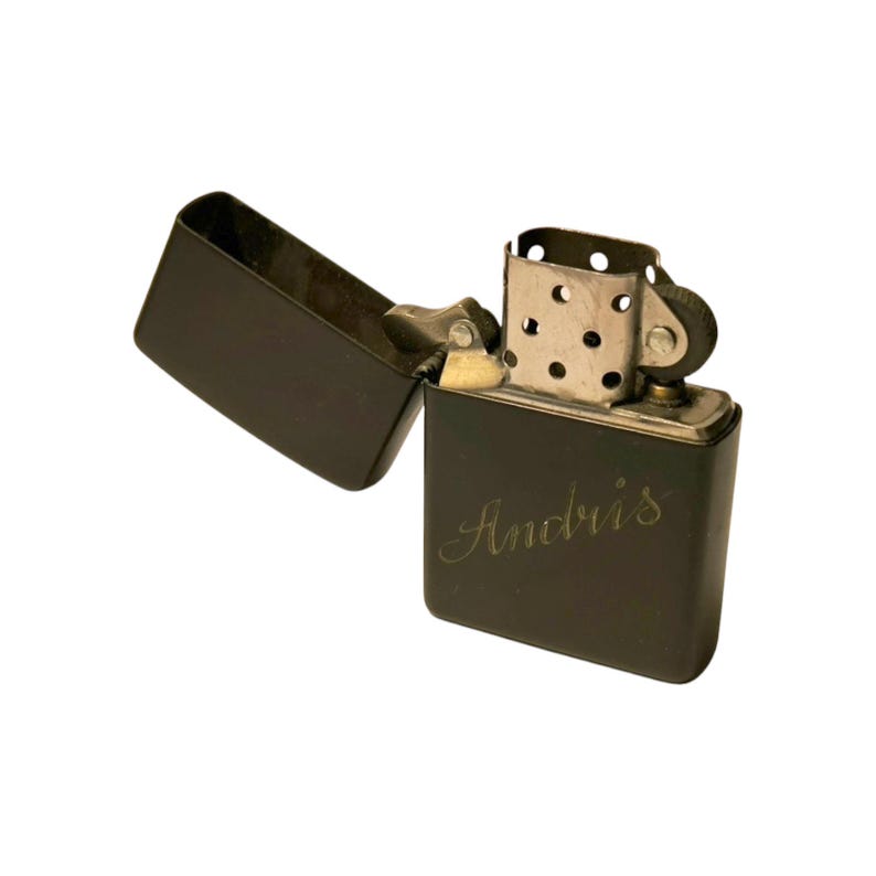 Puede incluir: Un encendedor de metal negro con la tapa abierta, revelando el mecanismo interno. El encendedor tiene el nombre "Andris" grabado en la parte delantera. El encendedor est&aacute; sobre un fondo blanco.