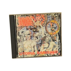 Op de afbeelding: Een cd-hoesje met de albumhoes van "Utopia Banished" van Napalm Death. De illustratie is een gedetailleerde, kleurrijke illustratie met een dystopisch thema, met figuren, tandwielen en het logo van de band.