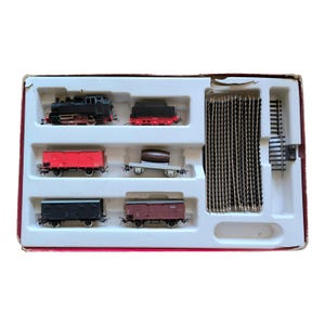 Puede incluir: Un set de tren modelo con una locomotora negra, un vagón rojo, un vagón marrón y un vagón plano gris con un barril. El set también incluye una sección de vía.