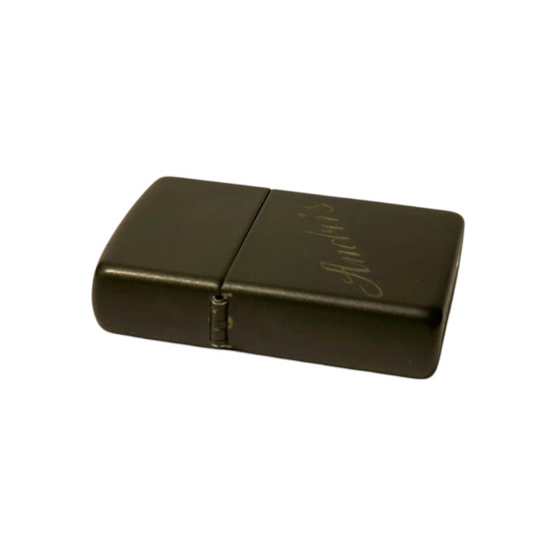 Puede incluir: Un encendedor de metal gris oscuro con la palabra "Andris" grabada en la parte delantera. El encendedor es rectangular con bordes redondeados y una tapa con bisagras. El encendedor est&aacute; cerrado en la imagen.