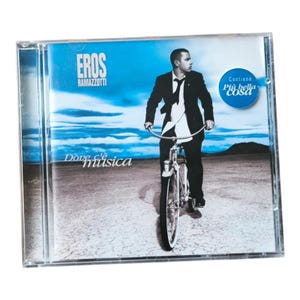 May include: A CD case featuring Eros Ramazzotti's album, "Dove c'è musica." The cover art shows Ramazzotti riding a bicycle, with a blue sky and desert landscape background. The CD case is in a clear plastic case.