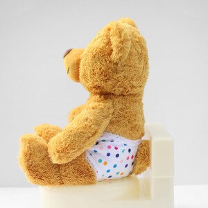 Puede incluir: Un oso de peluche marr&oacute;n con un pa&ntilde;al blanco con lunares multicolores est&aacute; sentado en un orinal blanco.