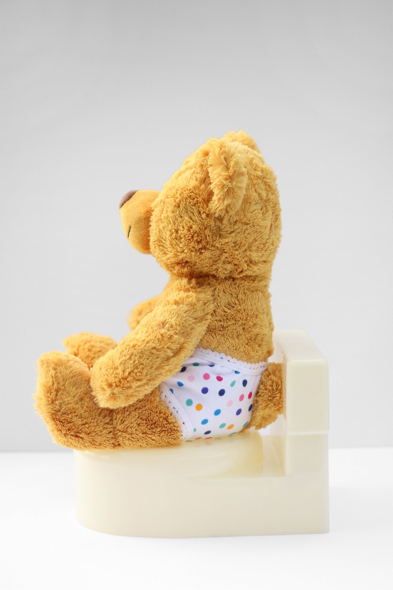Puede incluir: Un oso de peluche marr&oacute;n con un pa&ntilde;al blanco con lunares multicolores est&aacute; sentado en un orinal blanco.