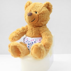 Puede incluir: Un oso de peluche marr&oacute;n con un pa&ntilde;al blanco con lunares de colores est&aacute; sentado en un orinal blanco.