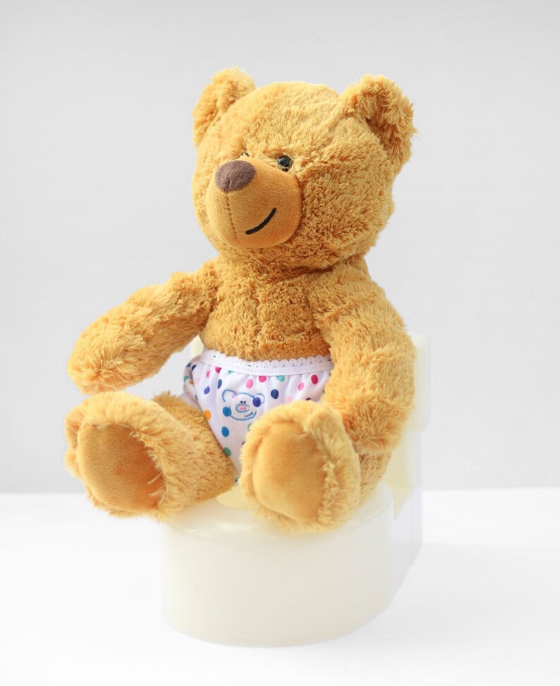Puede incluir: Un oso de peluche marr&oacute;n con un pa&ntilde;al blanco con lunares de colores est&aacute; sentado en un orinal blanco.