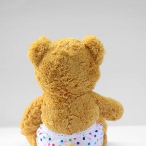 Puede incluir: Un oso de peluche marr&oacute;n que lleva un pa&ntilde;al blanco con lunares de colores.