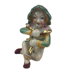 May include: A vintage porcelain figurine of a seated jester playing a trumpet. The jester wears a green and gold hooded hat, a colorful collar, and a matching outfit. The figurine has a white body, red shoes, and a gold trumpet.