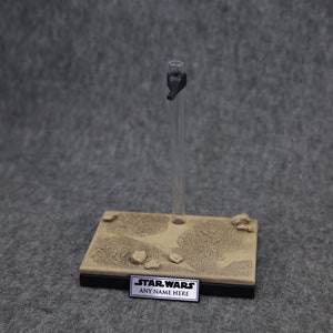 May include: A brown and black display stand with a clear plastic stand for a Star Wars action figure. The stand has a textured surface that resembles sand and has a black base with the text "STAR WARS ANY NAME HERE".