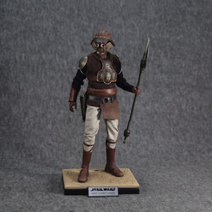 May include: A Star Wars action figure of a  Imperial Guard Lando, wearing a brown and gold armor with a helmet and holding a spear. The figure is standing on a brown base with the words "STAR WARS IMPERIAL GUARD LANDO" printed on it.