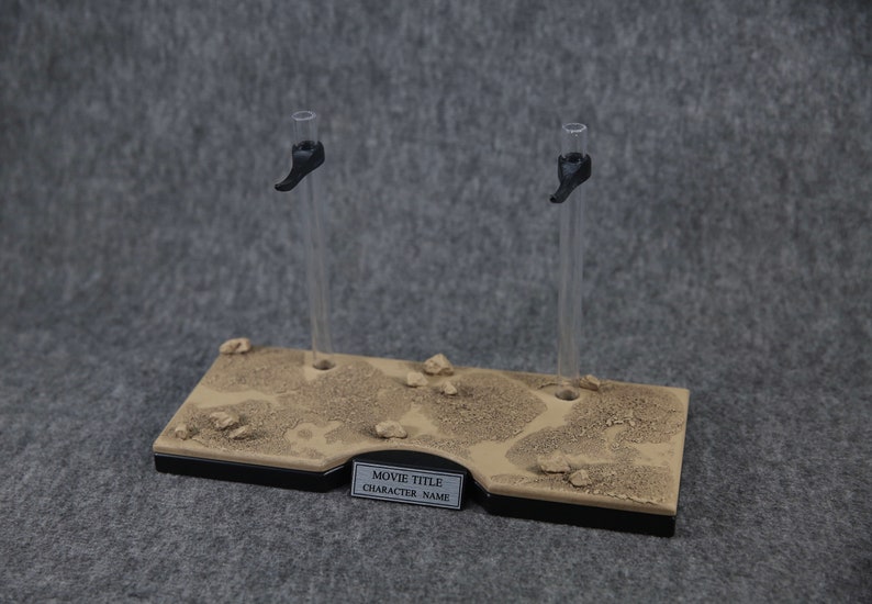 K&ouml;nnte beinhalten: Ein brauner Pr&auml;sentationsst&auml;nder mit zwei klaren Acryl-Stangen und schwarzen Clips. Der St&auml;nder hat eine W&uuml;sten-&auml;hnliche Textur und ein Etikett mit der Aufschrift "MOVIE TITLE CHARACTER NAME".