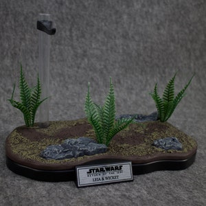 May include: A black display stand with a brown base and green ferns. The stand has a clear plastic rod and a black metal plate with the text "STAR WARS RETURN OF THE JEDI LEIA & WICKET".