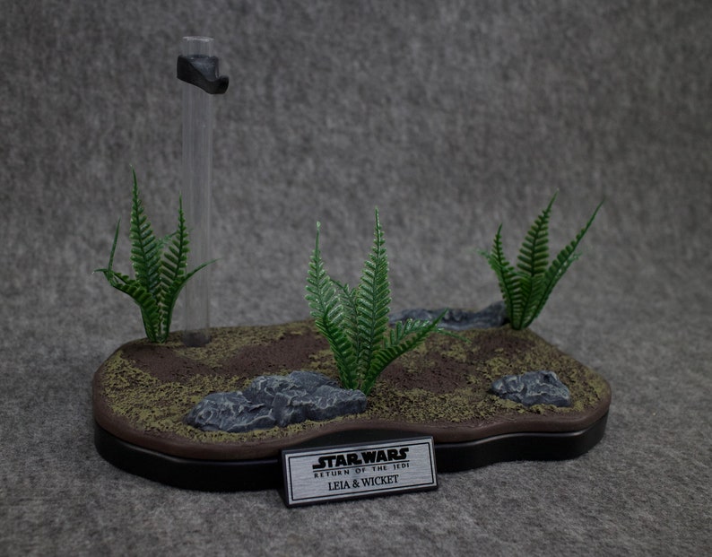 May include: A black display stand with a brown base and green ferns. The stand has a clear plastic rod and a black metal plate with the text "STAR WARS RETURN OF THE JEDI LEIA & WICKET".