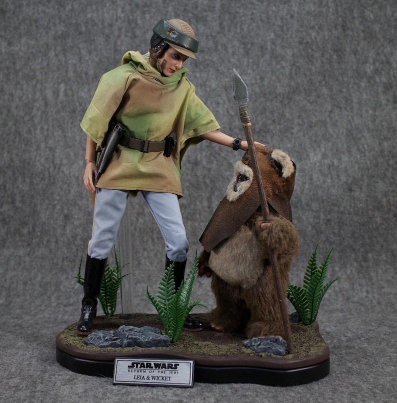 May include: A Star Wars action figure set featuring Princess Leia and Wicket the Ewok. Leia is wearing a green and brown poncho, white pants, and black boots. She is holding a blaster pistol. Wicket is wearing a brown fur coat and holding a wooden spear. The figures are standing on a black base with a green and brown landscape.