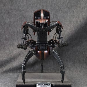 Puede incluir: Modelo detallado de un Droideka de Star Wars, un droide destructor rodante. La figura es principalmente negra y bronce, con extremidades articuladas y una cabeza detallada. La base presenta una etiqueta de Star Wars y Droideka.