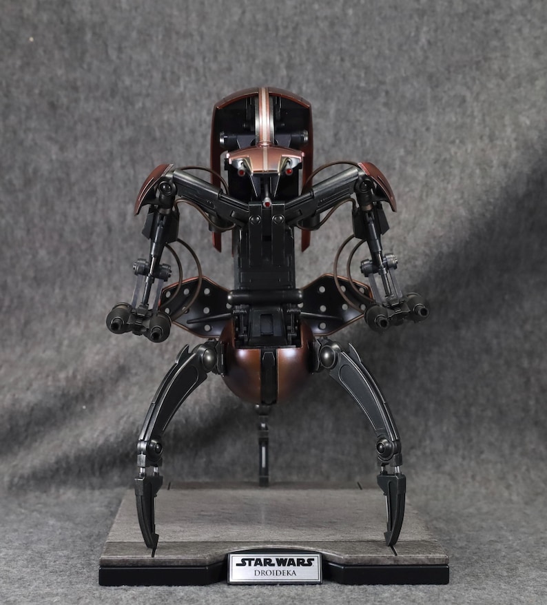 Puede incluir: Modelo detallado de un Droideka de Star Wars, un droide destructor rodante. La figura es principalmente negra y bronce, con extremidades articuladas y una cabeza detallada. La base presenta una etiqueta de Star Wars y Droideka.