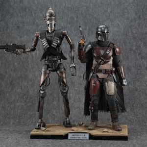 K&ouml;nnte beinhalten: Zwei Actionfiguren, eine eine metallische silberne und schwarze Roboterfigur mit einer Pistole, die andere eine schwarze und braune gepanzerte Figur mit einer Pistole, die auf einem braunen Sockel mit dem Text "MOVIE TITLE CHARACTER NAME" stehen.