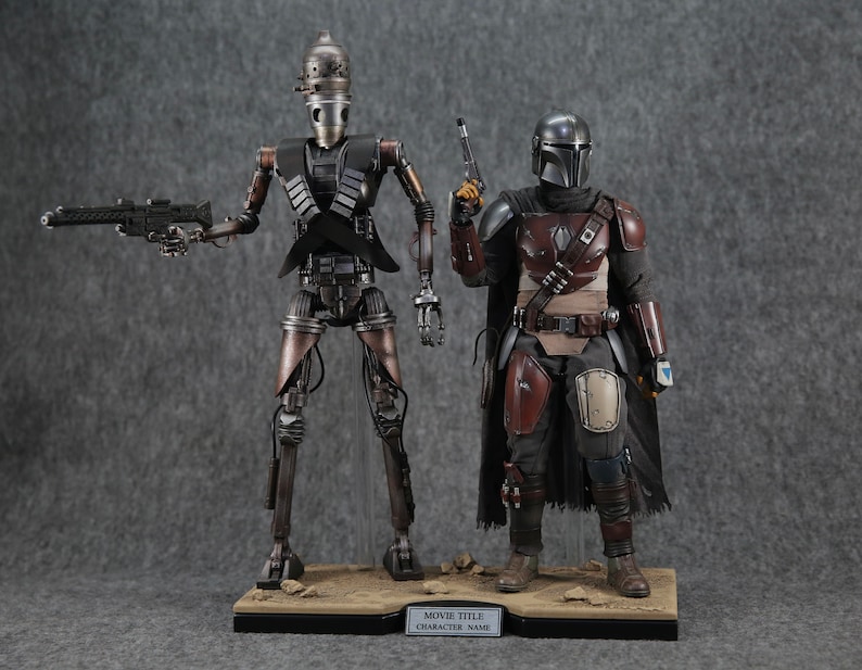 K&ouml;nnte beinhalten: Zwei Actionfiguren, eine eine metallische silberne und schwarze Roboterfigur mit einer Pistole, die andere eine schwarze und braune gepanzerte Figur mit einer Pistole, die auf einem braunen Sockel mit dem Text "MOVIE TITLE CHARACTER NAME" stehen.