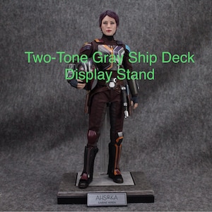 May include: A detailed action figure in a brown and orange outfit, holding a helmet. The figure stands on a gray display stand with the text "AHSOKA SABINE WREN". The text "Two-Tone Gray Ship Deck Display Stand" is also visible.