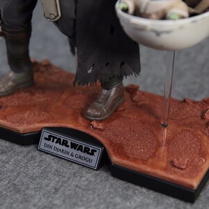 May include: A Star Wars action figure of Din Djarin, also known as The Mandalorian, holding a small, green alien character called Grogu, also known as Baby Yoda. The figures are standing on a brown, textured base with a black plaque that reads "Star Wars Din Djarin & Grogu".
