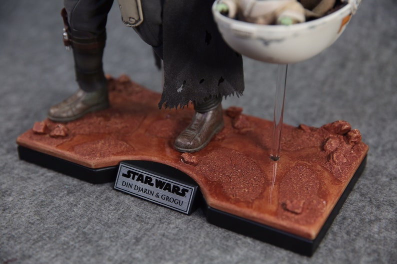 May include: A Star Wars action figure of Din Djarin, also known as The Mandalorian, holding a small, green alien character called Grogu, also known as Baby Yoda. The figures are standing on a brown, textured base with a black plaque that reads "Star Wars Din Djarin & Grogu".
