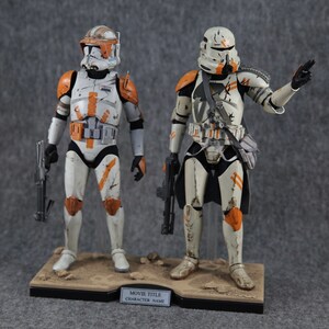 K&ouml;nnte beinhalten: Zwei Star Wars Actionfiguren, eine in orange-wei&szlig;er R&uuml;stung und die andere in wei&szlig;-schwarzer R&uuml;stung, beide mit verwitterter Farbe und Blastergewehren. Die Figuren stehen auf einem schwarzen Sockel mit einer braunen sandartigen Oberfl&auml;che.