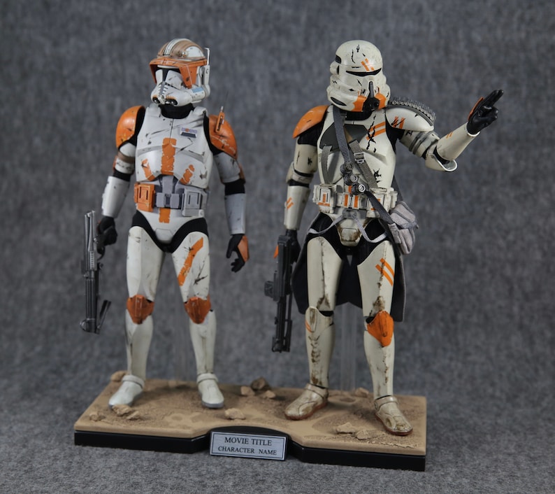 K&ouml;nnte beinhalten: Zwei Star Wars Actionfiguren, eine in orange-wei&szlig;er R&uuml;stung und die andere in wei&szlig;-schwarzer R&uuml;stung, beide mit verwitterter Farbe und Blastergewehren. Die Figuren stehen auf einem schwarzen Sockel mit einer braunen sandartigen Oberfl&auml;che.