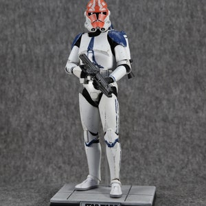 May include: A Star Wars 332nd Clone Trooper action figure. The figure is predominantly white with blue and orange accents. It is holding a black blaster rifle and stands on a gray base with the Star Wars logo and text.