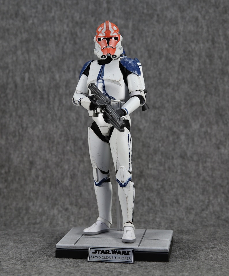 May include: A Star Wars 332nd Clone Trooper action figure. The figure is predominantly white with blue and orange accents. It is holding a black blaster rifle and stands on a gray base with the Star Wars logo and text.