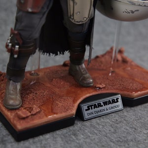 May include: Close-up of a Star Wars action figure of Din Djarin, also known as The Mandalorian, standing on a desert-themed display base. The figure is wearing a brown leather and metal armor with a black cape. The figure is holding a silver and white metal container. The base is made of brown plastic and has a silver plaque with the text "STAR WARS DIN DJARIN & GROGU".
