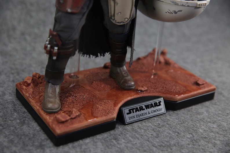 May include: Close-up of a Star Wars action figure of Din Djarin, also known as The Mandalorian, standing on a desert-themed display base. The figure is wearing a brown leather and metal armor with a black cape. The figure is holding a silver and white metal container. The base is made of brown plastic and has a silver plaque with the text "STAR WARS DIN DJARIN & GROGU".