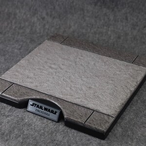 Puede incluir: Una base de exhibici&oacute;n rectangular con una superficie gris texturizada y un borde gris oscuro. La base presenta una peque&ntilde;a placa met&aacute;lica rectangular con el texto "STAR WARS DROIDKA".