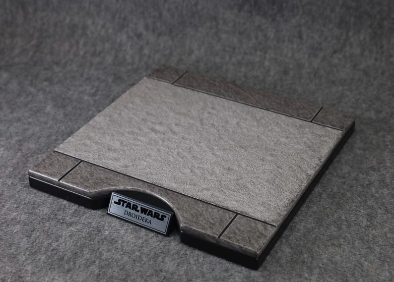 Puede incluir: Una base de exhibici&oacute;n rectangular con una superficie gris texturizada y un borde gris oscuro. La base presenta una peque&ntilde;a placa met&aacute;lica rectangular con el texto "STAR WARS DROIDKA".