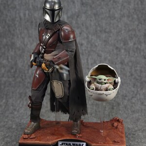 May include: A Star Wars action figure set featuring Din Djarin, also known as The Mandalorian, and Grogu, also known as Baby Yoda. The Mandalorian is wearing his signature brown and black armor and holding a blaster rifle. Grogu is sitting in a floating pod. The figures are displayed on a desert-themed base.