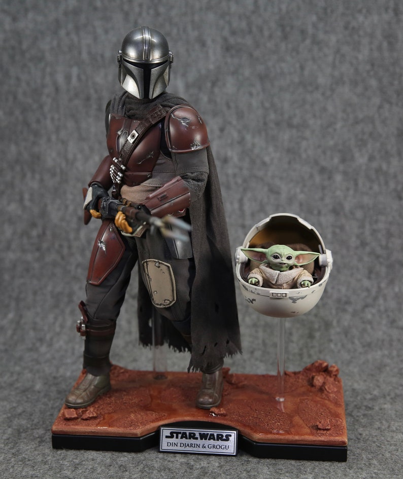 May include: A Star Wars action figure set featuring Din Djarin, also known as The Mandalorian, and Grogu, also known as Baby Yoda. The Mandalorian is wearing his signature brown and black armor and holding a blaster rifle. Grogu is sitting in a floating pod. The figures are displayed on a desert-themed base.
