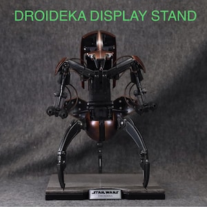 Puede incluir: Un soporte de exhibici&oacute;n detallado para una figura de Droideka, con una combinaci&oacute;n de colores marr&oacute;n y negro met&aacute;lico. El soporte tiene un logotipo de Star Wars y el texto "DROIDEKA DISPLAY STAND" en verde.
