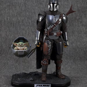 Puede incluir: Un conjunto de figuras de acci&oacute;n detallado con The Mandalorian con armadura plateada y negra, con capa y bl&aacute;ster. Incluye una figura de Grogu en una c&aacute;psula flotante. La base tiene el texto "STAR WARS DIN DJARIN & GROGU".
