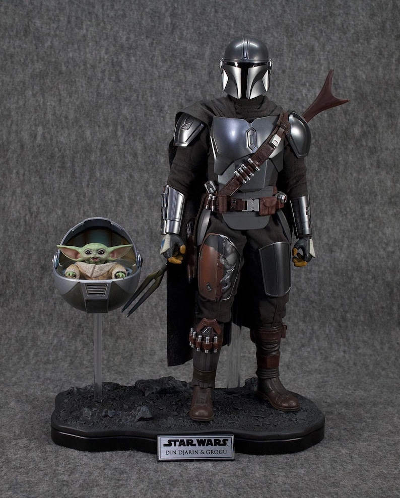 Puede incluir: Un conjunto de figuras de acci&oacute;n detallado con The Mandalorian con armadura plateada y negra, con capa y bl&aacute;ster. Incluye una figura de Grogu en una c&aacute;psula flotante. La base tiene el texto "STAR WARS DIN DJARIN & GROGU".